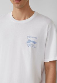Weißes Baumwoll-T-Shirt mit kurzen Ärmeln. Enthält einen kleinen blauen Grafikdruck auf der linken Brust mit der Aufschrift "LAZY sunny Pool Days." Glatte Textur.