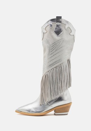Botte de cowboy en argent avec un bout pointu, des franges décoratives et des strass sur une tige en daim. Présente un talon en bois empilé.