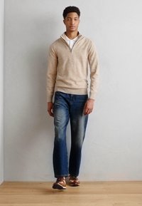 Beige kötött pulóver, cipzáros nyakkal, kék farmernadrággal párosítva. Barna és fehér tornacipők egészítik ki az öltözéket. Egyszerű háttér.