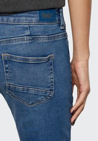 Jeans de denim azuis com um corte slim, apresentando um bolso traseiro e uma etiqueta de marca na cintura. A cor é azul média com costuras claras.