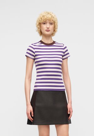 Γυναίκα με ξανθά σγουρά μαλλιά, ντυμένη με εφαρμοστό μπλουζάκι με κοντά μανίκια σε μωβ και λευκές ρίγες και μαύρη φούστα σε σχήμα Α, στέκεται μπροστά σε απλό φόντο.