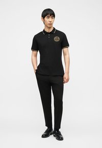 Άνδρας μοντέλο στέκεται φορώντας μαύρο πόλο πουκάμισο με χρυσό λογότυπο και περιθώριο, μαύρο παντελόνι και μαύρα επίσημα παπούτσια μπροστά σε μονόχρωμο φόντο.