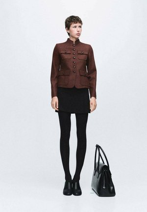 Mujer con chaqueta marrón abotonada, falda negra, mallas negras y botines de tobillo, de pie junto a un bolso negro sobre un fondo liso.
