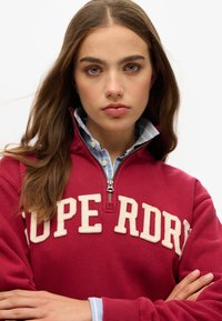 Bordeaux zip-up sweatshirt met crèmekleurige geborduurde letters "SUPERDRY" op de borst; gelaagd over een lichtblauw, geruit overhemd met kraag.