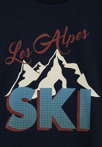 Námořnický svetr s bílou grafikou hor, červeným nápisem "Les Alpes" a velkým nápisem "SKI" v modrých a zelených puntících, s červenými akcenty.