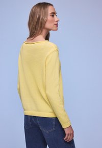 Femme aux cheveux longs et blonds, portant un pull en maille jaune et un jean bleu foncé, légèrement tournée de côté sur un fond bleu pâle.