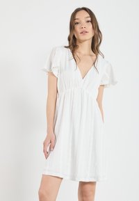 Valkoinen mekko, jossa on V-pääntie, lyhyet liehuvat hihat, rakenteellinen kangas ja rypytetty vyötärö. Mekko ylettyy polvien ylle ja on rento malli.