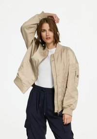 Beige Bomberjacke mit gerippten Bündchen und Kragen, mit Reißverschluss und Seitentaschen, kombiniert mit dunkelhosen. Glatte Textur.