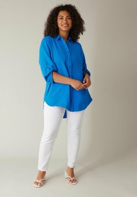 Camicia azzurra brillante con maniche arrotolate, abbinata a pantaloni bianchi slim-fit e sandali bianchi con cinturini. Texture liscia, design casual.