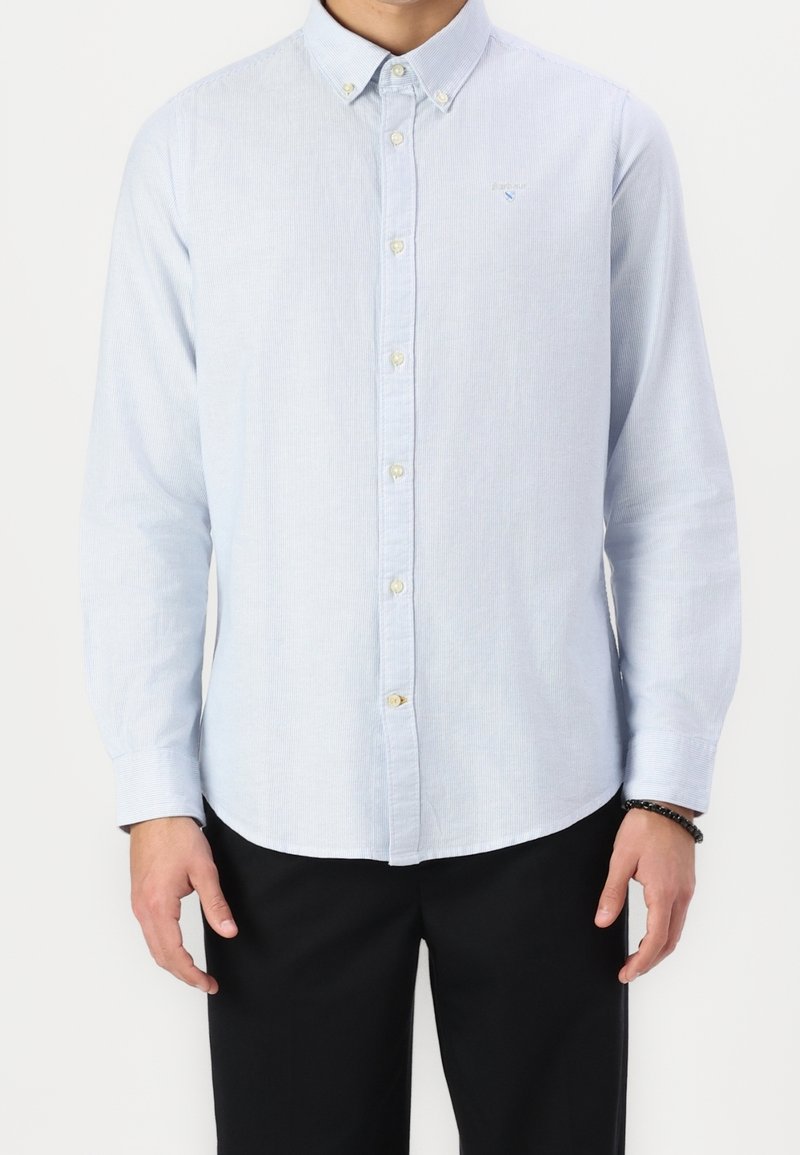 Homme portant une chemise à rayures bleu clair à manches longues, rentrée dans un pantalon noir, avec un bracelet de perles noires au poignet droit.