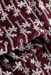 Tissu bordeaux avec une broderie florale blanche complexe disposée en lignes verticales, montrant la texture et les plis du tissu.