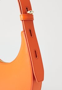 Oranje leren handtas met een ronde vorm, voorzien van een platte schouderriem met gaatjes en een gouden accent. Zachte textuur en minimalistisch ontwerp.