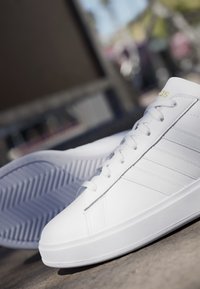 Hvid snøresneaker med tekstureret sål og tre diskrete sidelommer placeret på en betonoverflade med sløret udendørs baggrund.