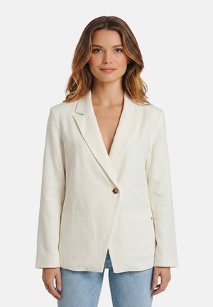 Mujer con cabello ondulado hasta los hombros, vistiendo un blazer crema con un solo botón y unos vaqueros azul claro, de pie frente a un fondo liso.
