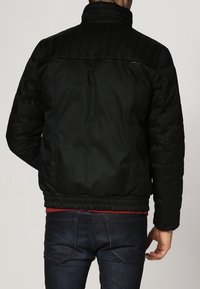 Chaqueta acolchada negra con cuello alto, cintura elástica y puños de canalé. Presenta un detalle de costuras horizontales en los hombros y la espalda.