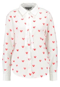 Vit blus med ett mönster av röda hjärtan, standardkrage, långa ärmar, knäppning framtill och en knytning vid halsen.