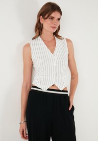 Bela brezrokavnik s gumbi in tankimi črnimi vertikalnimi črtami, z V-izrezom in skrajšanim krojem, v kombinaciji s črnimi hlačami z visokim pasom.