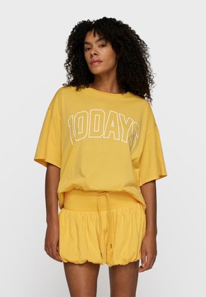 Kobieta o kręconych włosach, ubrana w luźną żółtą koszulkę z napisem "10 DAYS" i pasujące żółte szorty z marszczeniem, na jednolitym tle.