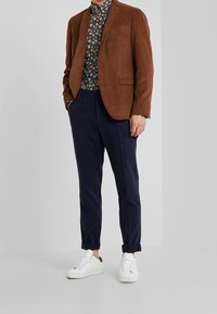Brun kordflodig blazer över en marinblå mönstrad skjorta, i kombination med marinblå byxor och vita sneakers. Modern passform med upprullade ärmar och synliga fickor.