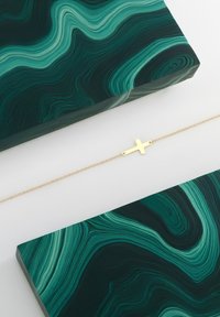 Goldenes Kreuzarmband mit einer zarten Kette, platziert auf einem dunklen grünen, marmorierten Hintergrund mit fließenden Mustern und verschiedenen Grüntönen.