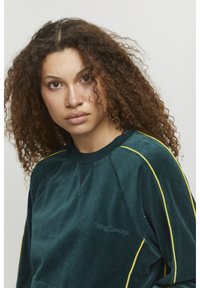 Felpa in velluto verde scuro con strisce laterali gialle, dotata di collo tondo, polsini a coste e un piccolo logo ricamato sul petto.