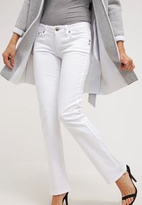 Pantalones vaqueros blancos de corte ajustado, diseño de cinco bolsillos y detalles de costura sutiles, complementados con sandalias negras de tacón.