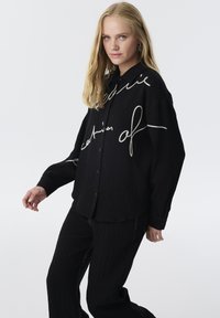 Schwarzes Langarmshirt aus strukturiertem Stoff, mit weißer gestickter Schrift. Kombiniert mit schwarzen Hosen, die kontrastierende Nähte aufweisen.