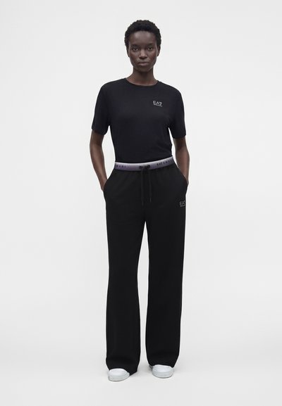Жена стои, облечена в черна тениска EA7 и черен панталон с широки крачоли и емблематична талия, ръце в джобовете, бели маратонки, обикновен фон.