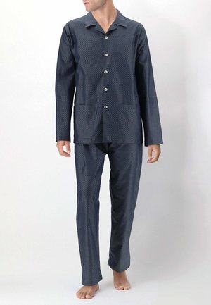 Uomo che indossa un pigiama blu scuro a maniche lunghe con bottoni e un piccolo motivo a pois, in piedi a piedi nudi su uno sfondo bianco uniforme.