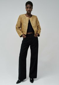 Chaqueta de lentejuelas doradas con escote redondo, combinada con pantalones anchos negros y zapatos de punta negra, sobre un fondo neutro.