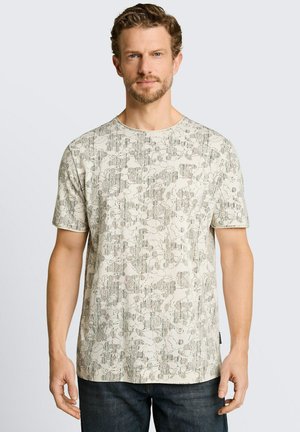 Man met kort krullend haar en baard, gekleed in een beige T-shirt met patroon en korte mouwen en een donkere broek, staand voor een eenvoudige lichte achtergrond.