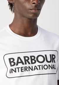Detailní záběr na osobu v bílé tričku s tučným černým textem "BARBOUR INTERNATIONAL" v obdélníkovém rámečku.