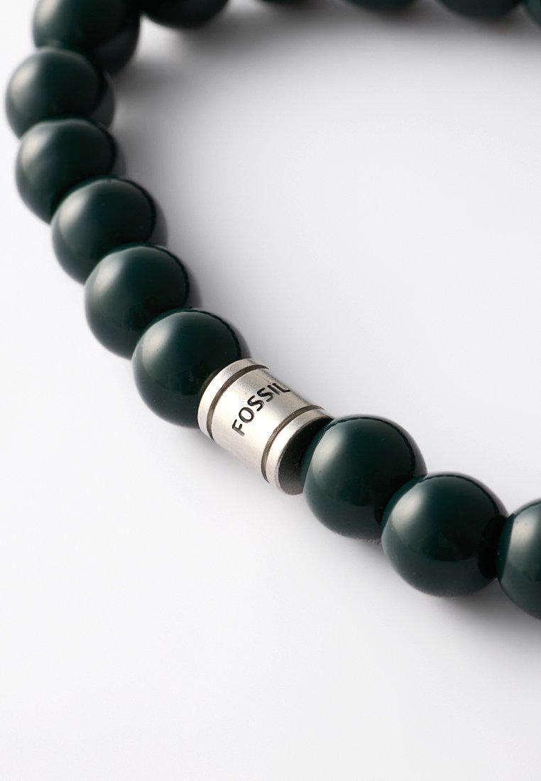 Bracelet en perles vert foncé, comprenant des pierres rondes brillantes et un accent en argent avec "FOSSIL" gravé. Texture lisse, design élastique.