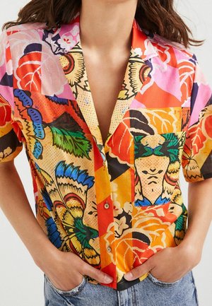 Vrouw met een gebloemd kort mouw shirt met knopen, met levendige multicolor bloemen- en abstracte patronen, handen gestoken in de zakken van lichtblauwe spijkerbroeken.