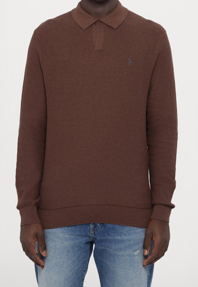 Homme portant un pull polo marron à manches longues avec des poignets côtelés et un jean bleu clair sur un fond uni.