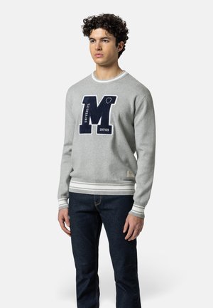 Jonge man draagt een grijze trui met een grote marineblauwe "M" en de tekst "UNIVERSITÀ 19769," gecombineerd met donkerblauwe spijkerbroek, staand tegen een witte achtergrond.