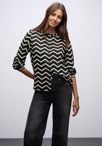 Blusa de chevron en negro y crema con botones, cuello redondo y mangas tres cuartos, combinada con jeans oscuros. Textura suave.