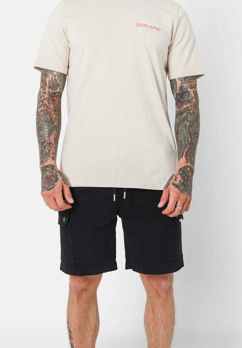 T-shirt in cotone beige con logo rosa sul petto, abbinato a pantaloni cargo neri con tasche laterali e vita regolabile con cordino.
