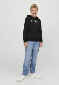 Sudadera con capucha negra y logo blanco, combinada con jeans de color azul claro y zapatillas blancas, diseñada para el uso casual juvenil.