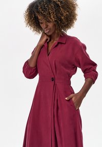 Kobieta o kręconych włosach, ubrana w długą, bordową sukienkę na guziki z długim rękawem, jedna ręka w kieszeni, druga delikatnie dotykająca szyi.