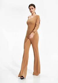 Eén-schouder, aansluitende jumpsuit in tan stof. Lange mouw met zij uitsnede, flared broekspijpen, gladde textuur, geen zichtbare patronen of hardware.