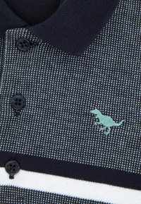 Marinblå stickad polo med ett texturerat mönster, tre svarta knappar och en mintgrön broderad dinosaurie-logotyp på vänster bröst.