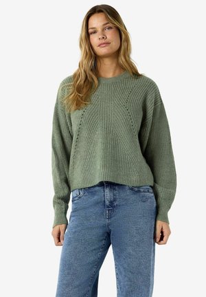 Ung kvinde med langt blond hår iført en løs grøn striktrøje og blå jeans, stående mod en ensfarvet hvid baggrund.