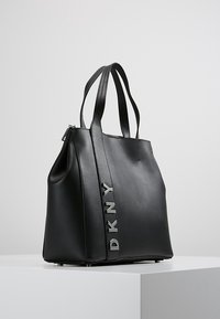 Czarna skórzana torba na ramię z podwójnymi uchwytami, wyraźne logo DKNY w kolorze srebrnym z boku, zdefiniowany kształt, gładka tekstura i płaska podstawa.
