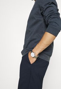 Braccio che indossa un orologio da polso in argento con un quadrante bianco, mano in tasca, vestito con una camicia a maniche lunghe blu scuro e pantaloni blu navy.