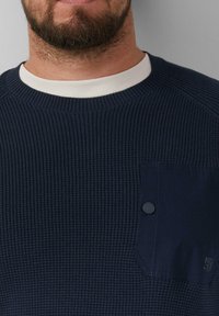 Marineblauer gerippter Pullover mit einem weißen Kragen, ausgestattet mit einer knöpfbaren Taschenapplikation auf der Brust und strukturiertem Stoff für zusätzliche Tiefe.
