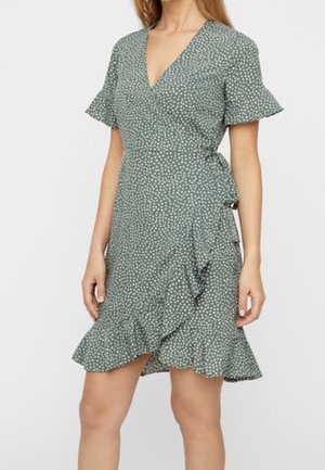 Vestido envolvente verde con lunares blancos, mangas cortas acampanadas, escote en V y volante en el dobladillo, llevado por una persona de pie.