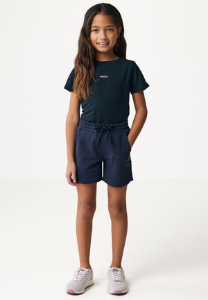 T-shirt bleu marine foncé avec un petit logo rose, associé à un short bleu marine assorti avec une taille élastique et des poches latérales. Baskets légères.