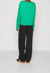 Sudadera verde de gran tamaño con cuello redondo, combinada con pantalones cargo negros que cuentan con múltiples bolsillos y un ajuste relajado.
