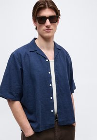 Άνδρας με σκούρα γυαλιά ηλίου, μπλε ναυτικό κοντομάνικο πουκάμισο με κουμπιά πάνω από λευκό αμάνικο μπλουζάκι, με καφέ παντελόνια και τα χέρια στις τσέπες.